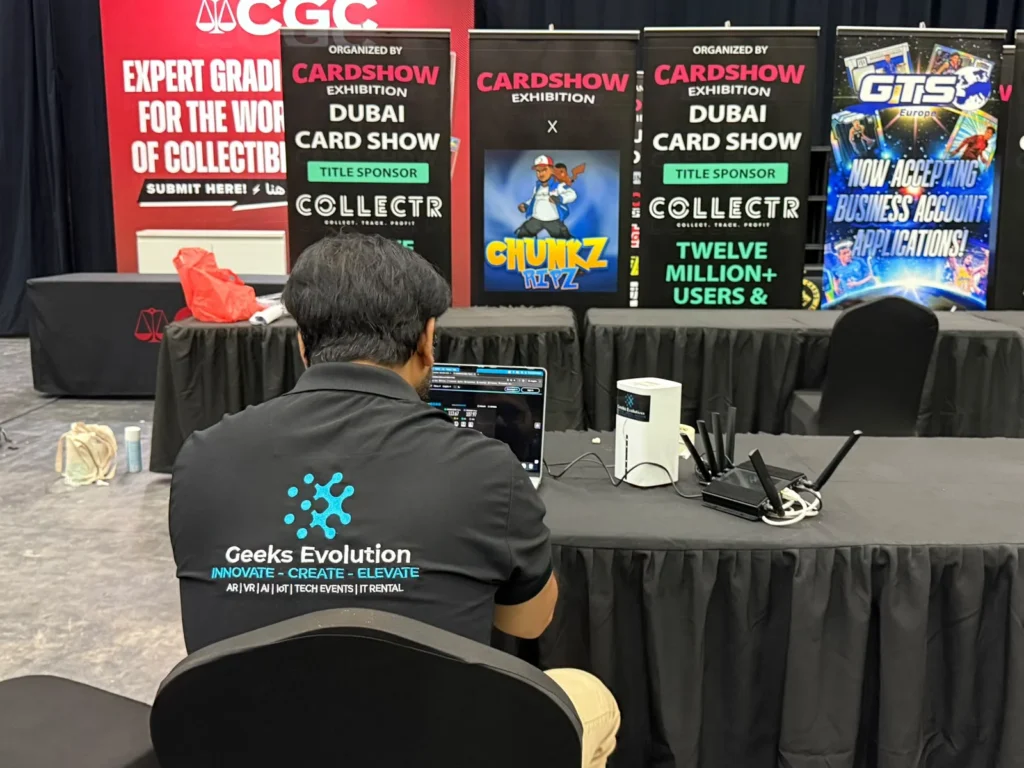 Geeks Evolution engineer testing event internet connectivity on a laptop beside network routers at a Dubai card show exhibition.